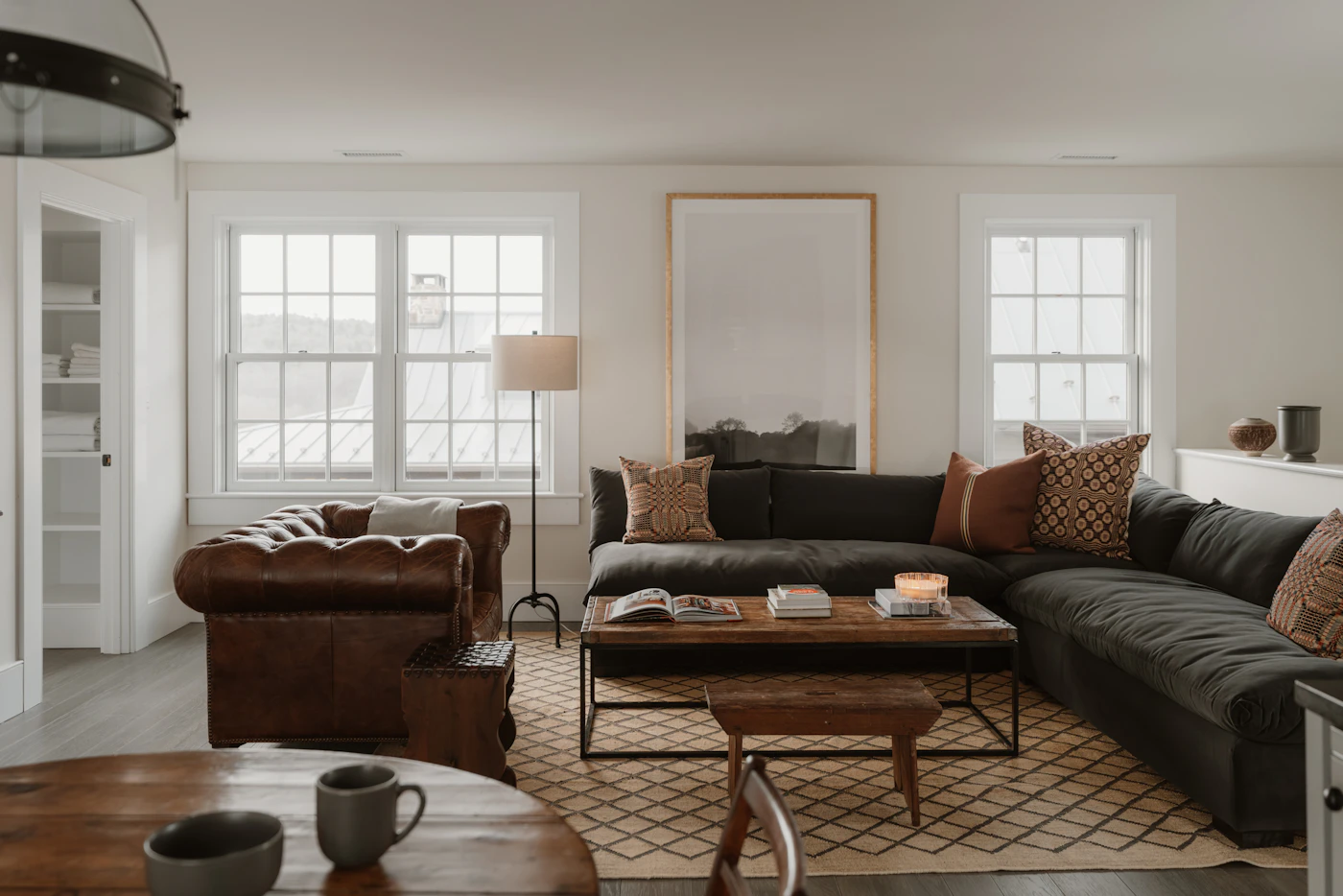 Dark sectional and brown chair beside a shaded floor lamp and timber coffee table in a bright living room.